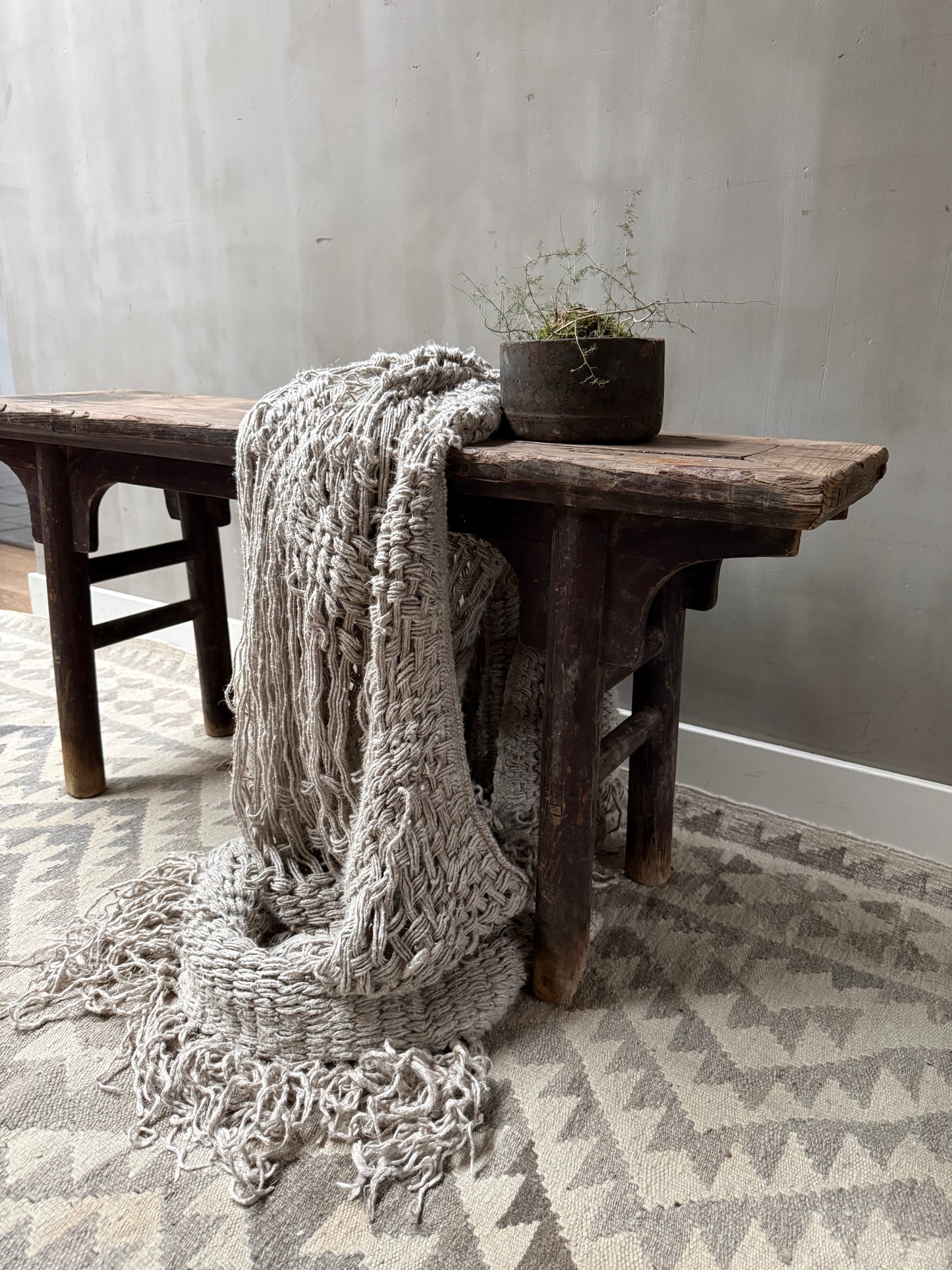 Landelijk houten bankje met grof gebreid plaid en zwarte pot met plant, sober interieur