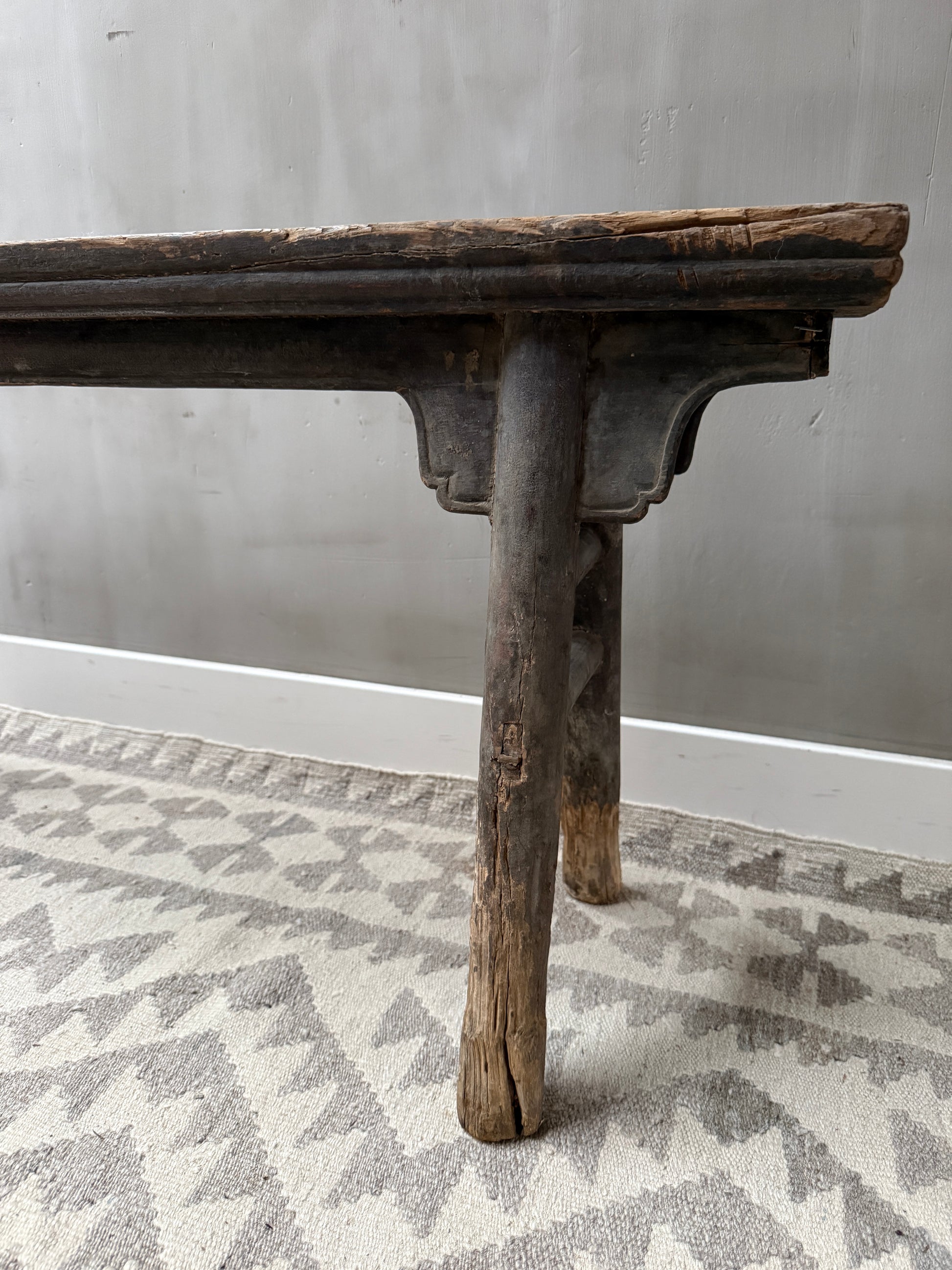 Authentiek handgemaakt houten bankje met verweerde poten op een licht vloerkleed, sober landelijk interieur