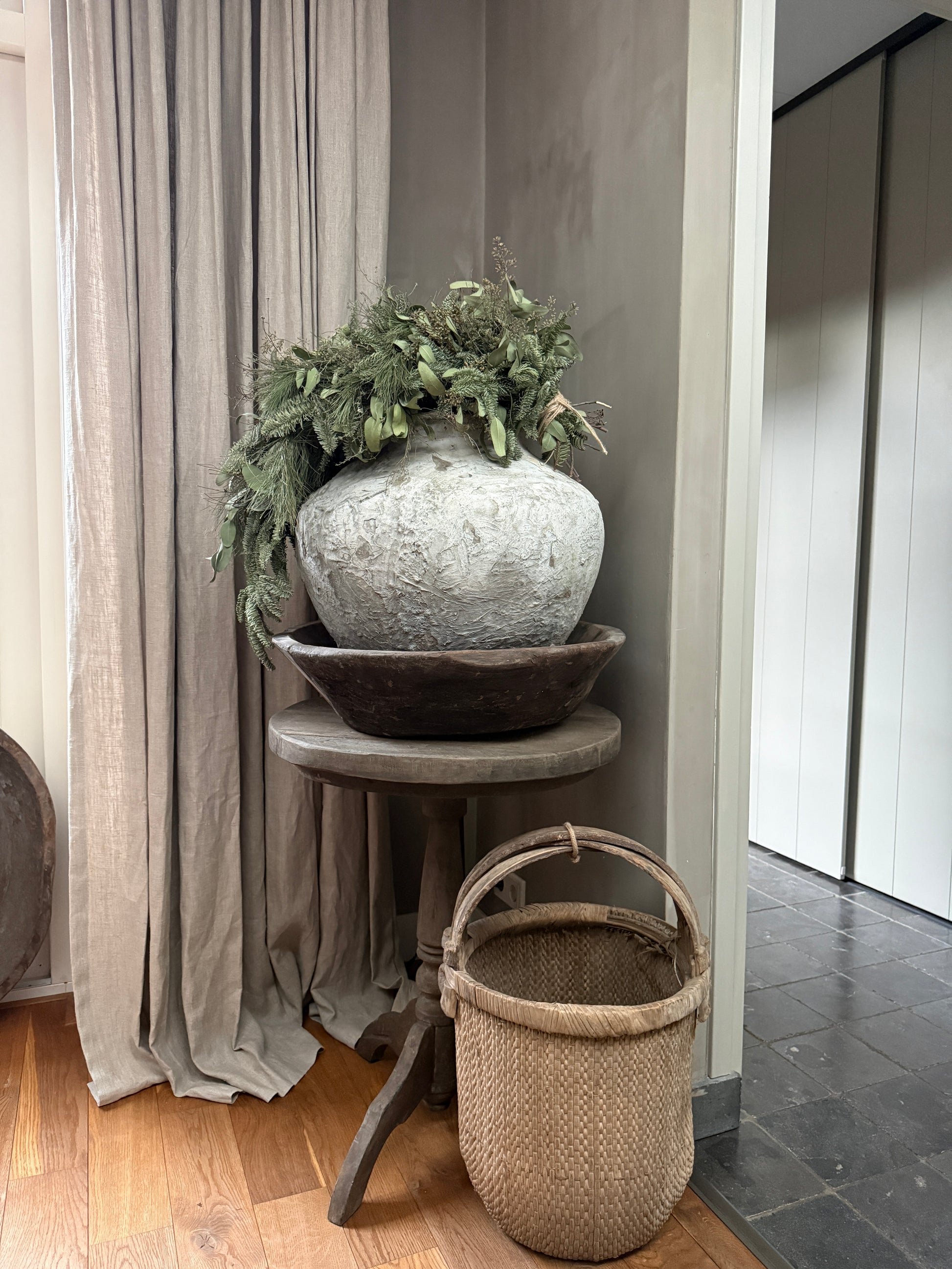 Handgemaakte landelijke vaas met groen op oud houten tafel, rieten mand ernaast, sober interieur