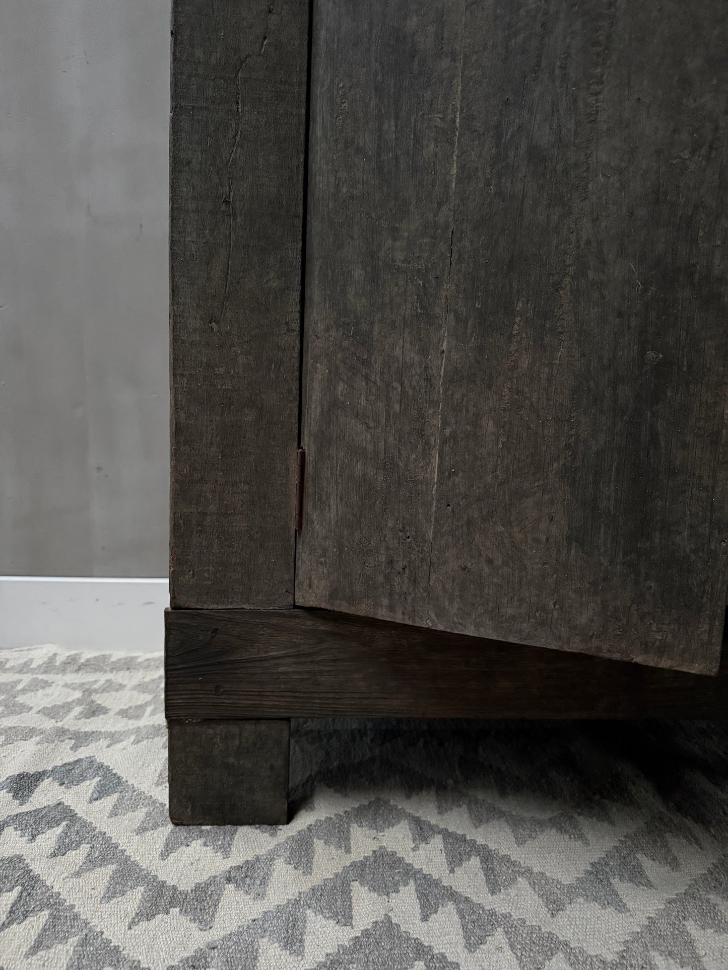 Close-up van een handgemaakte houten kastpoot op een grijs-wit patroon vloerkleed, landelijk en sober interieur.