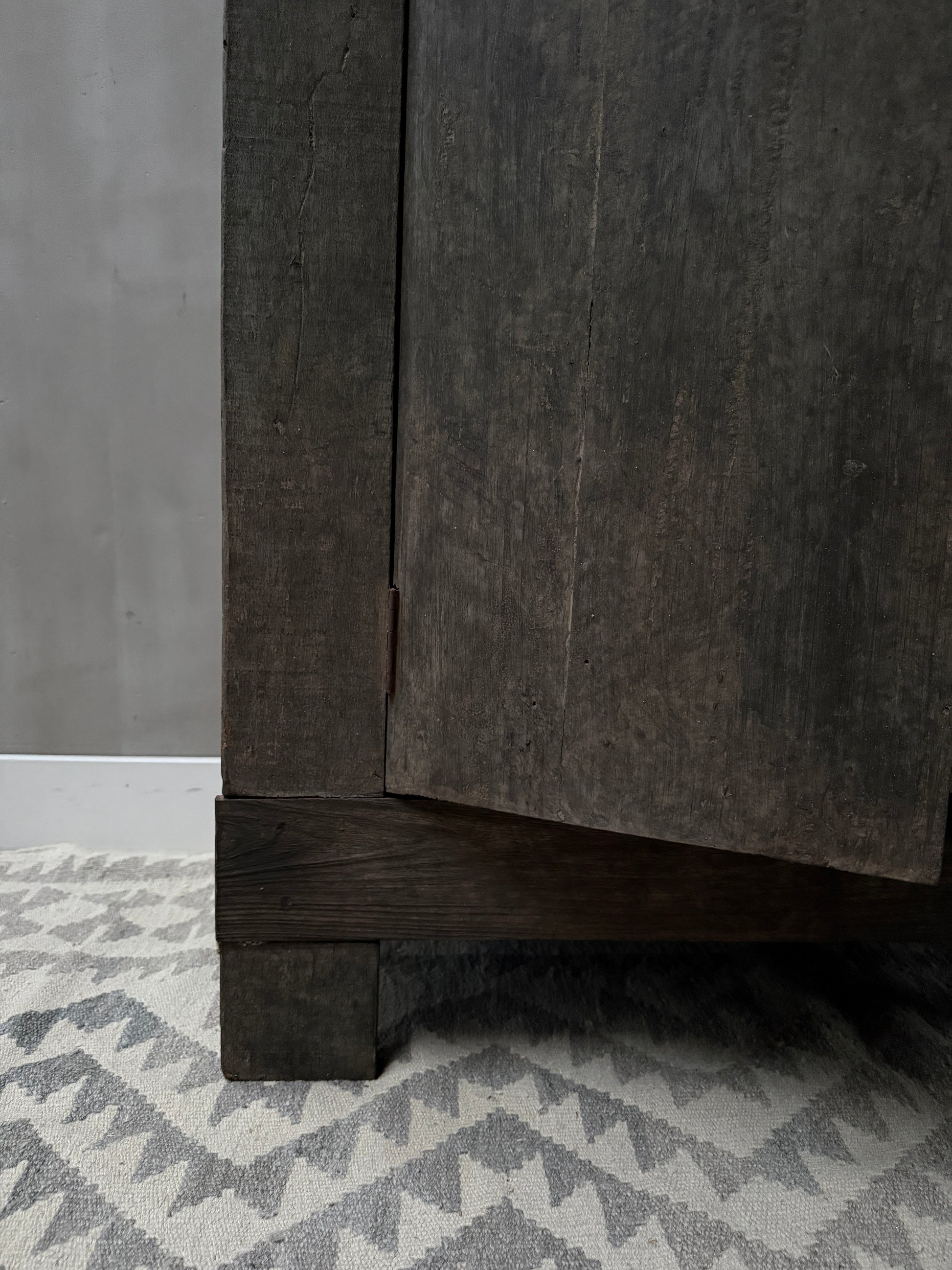 Close-up van een handgemaakte houten kastpoot op een grijs-wit patroon vloerkleed, landelijk en sober interieur.