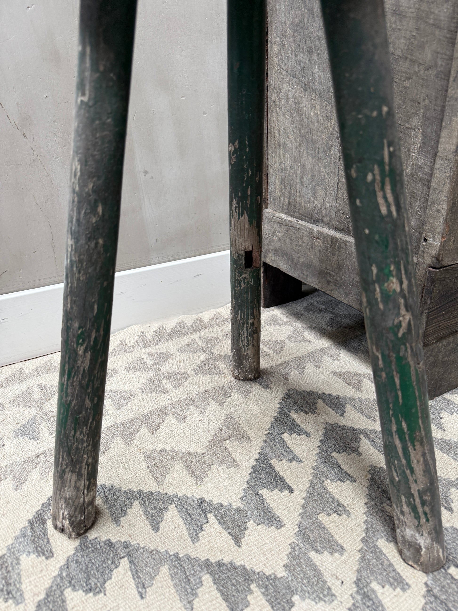Authentieke, verweerde houten kruk met groene poten op beige vloerkleed met patroon, sober interieur