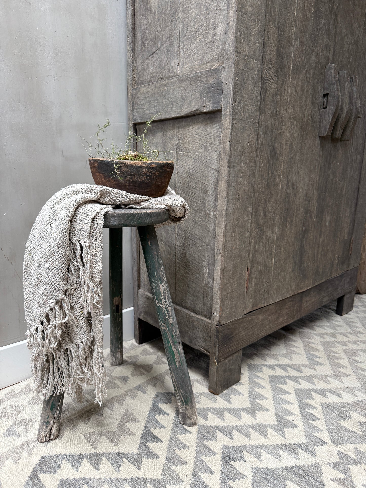 Landelijke houten kruk met grijze plaid en handgemaakt schaaltje bij sober dressoir op vloerkleed