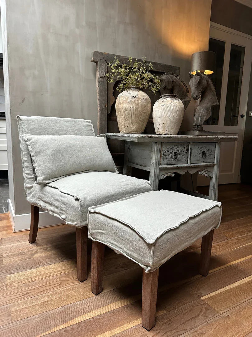 Landelijke fauteuil met linnen bekleding en kruk, naast oude houten tafel met keramische vazen