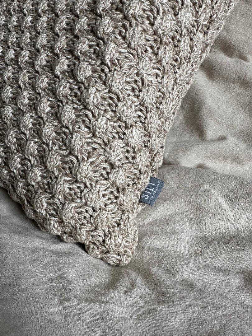 Handgemaakt grof gebreid kussen in beige tinten op linnen beddengoed, landelijk interieur