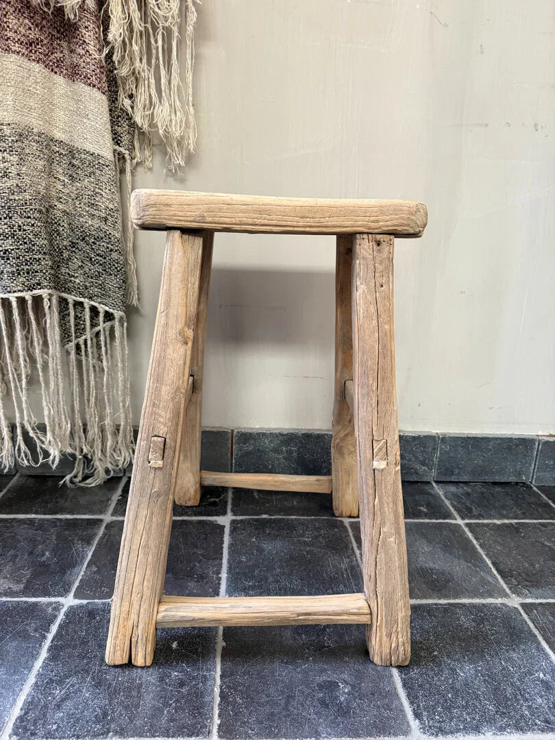 Authentiek handgemaakt houten krukje op zwarte tegelvloer, sober landelijk interieur