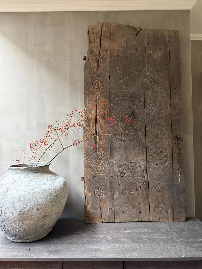 Handgemaakt houten paneel met etnisch motief, grote sobere vaas met rode takken op tafel, landelijk interieur