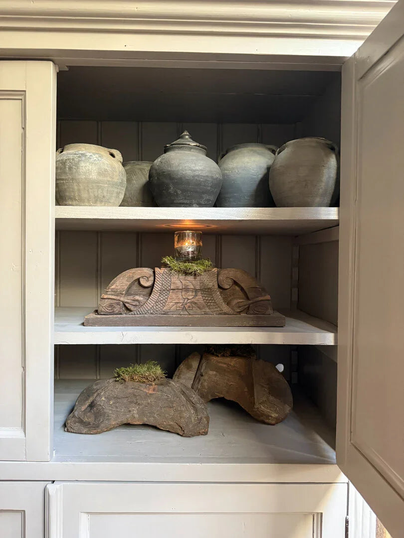 Houten kandelaar met glas en mos op plank, omringd door vintage potten in sobere landelijke kast