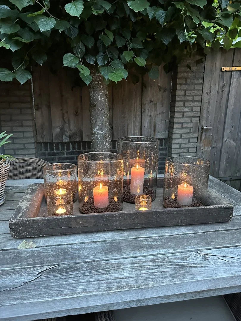 Windlicht van glas met steenslag en brandende kaarsen op een houten schaal, landelijke sfeer decoratie.
